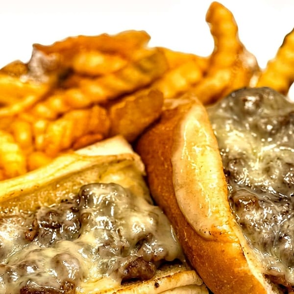 Two cheesy steak sandwiches with crinkle-cut fries on a white background.